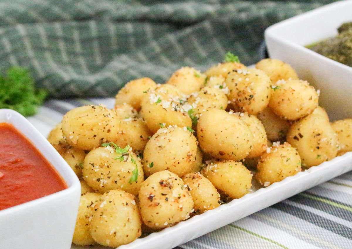 A rectangular white plate with seasoned, bite-sized potato balls garnished with herbs, accompanied by a square dish of red dipping sauce. A green checkered cloth is visible in the background.