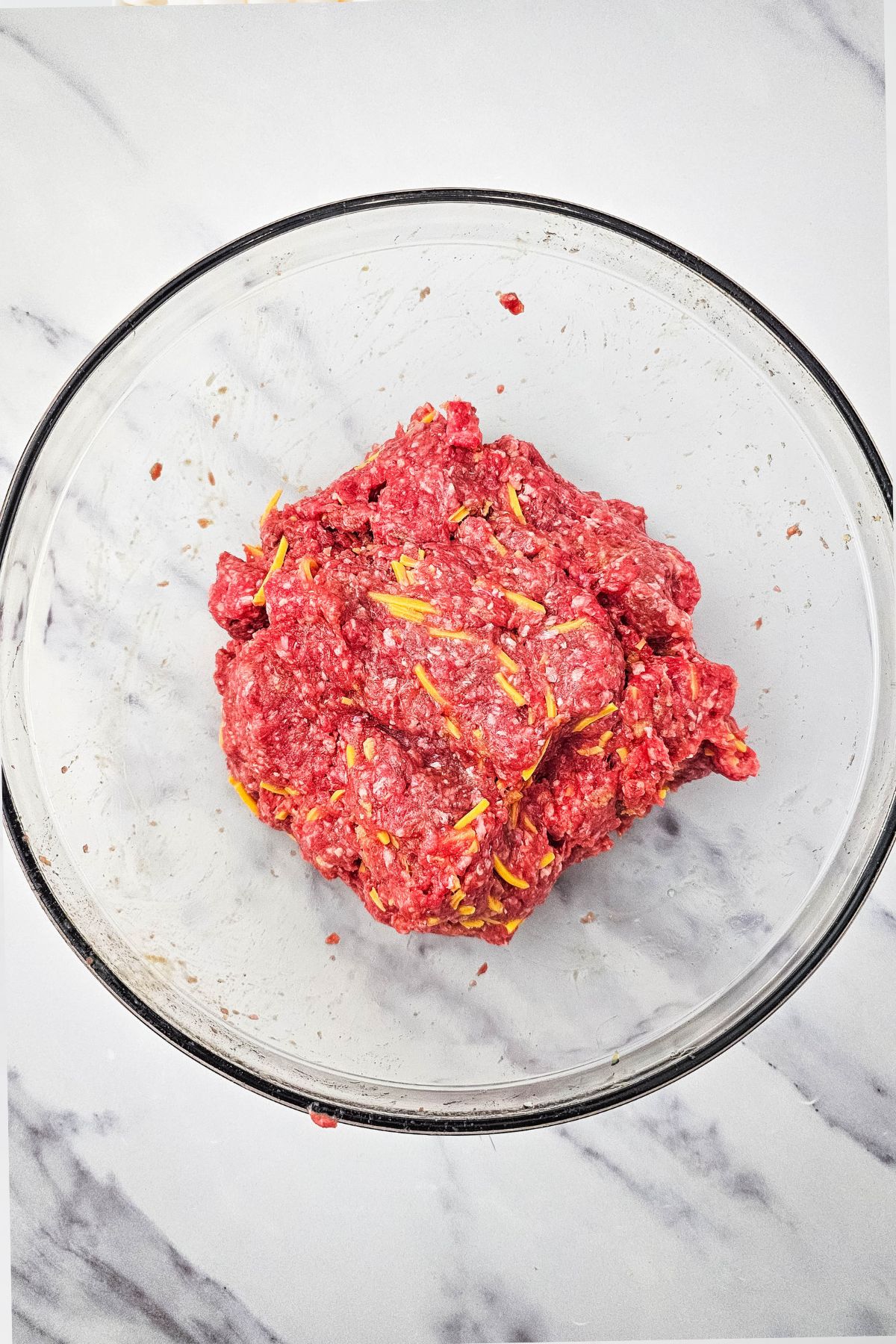 A clear glass bowl containing a mixture of raw ground beef and shredded cheese sits on a white marble surface.