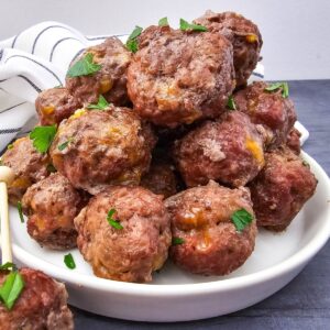 A white plate is piled with browned, oven-baked meatballs garnished with chopped parsley. A white dish towel with black stripes is visible in the background on a dark surface.