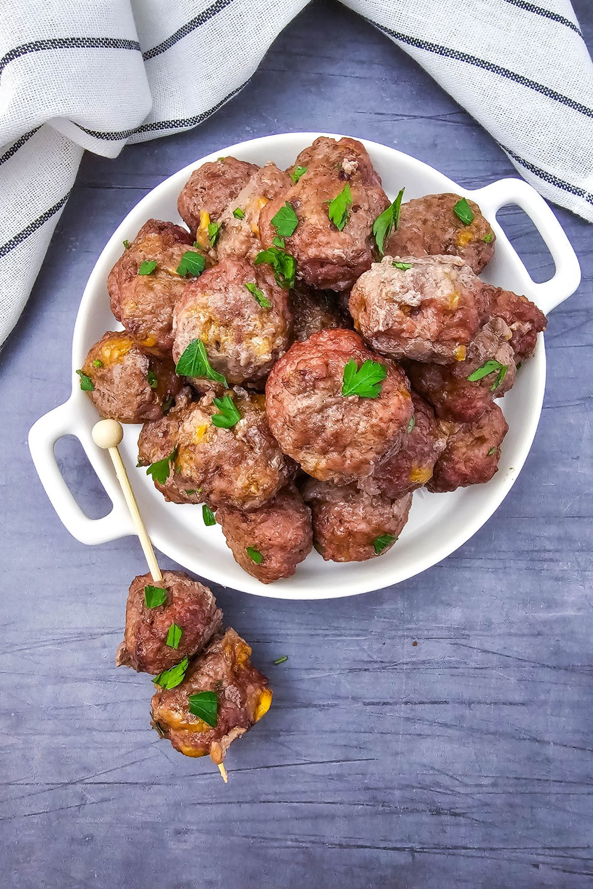 A white plate filled with several cooked meatballs garnished with chopped parsley, with a wooden skewer holding two meatballs resting next to the plate, on a gray surface beside a folded striped cloth.