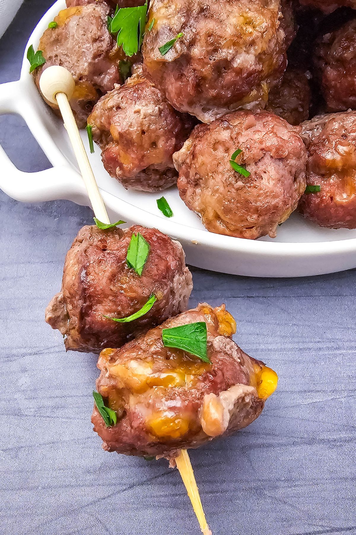 Two meatballs on a wooden skewer rest in front of a white dish filled with more meatballs. The meatballs contain melted cheese and are garnished with chopped parsley. The background surface is gray.
