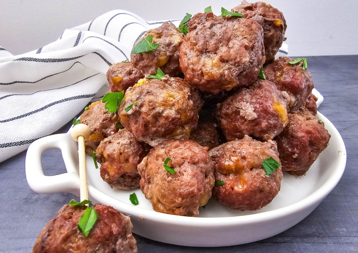 A plate stacked with several cooked meatballs garnished with chopped parsley, with one meatball on a skewer in the foreground. A white cloth with black stripes is in the background.