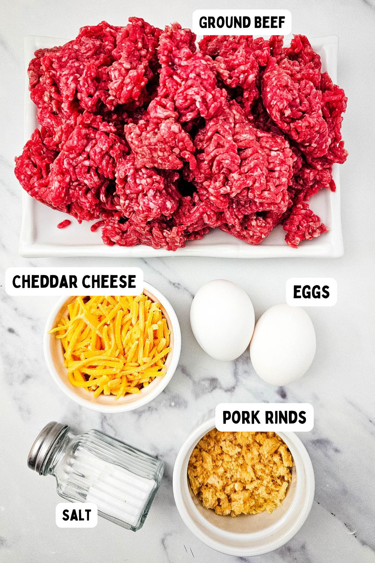 A white surface displays labeled ingredients: ground beef on a rectangular plate, shredded cheddar cheese in a bowl, two whole eggs, crushed pork rinds in a bowl, and a salt shaker.