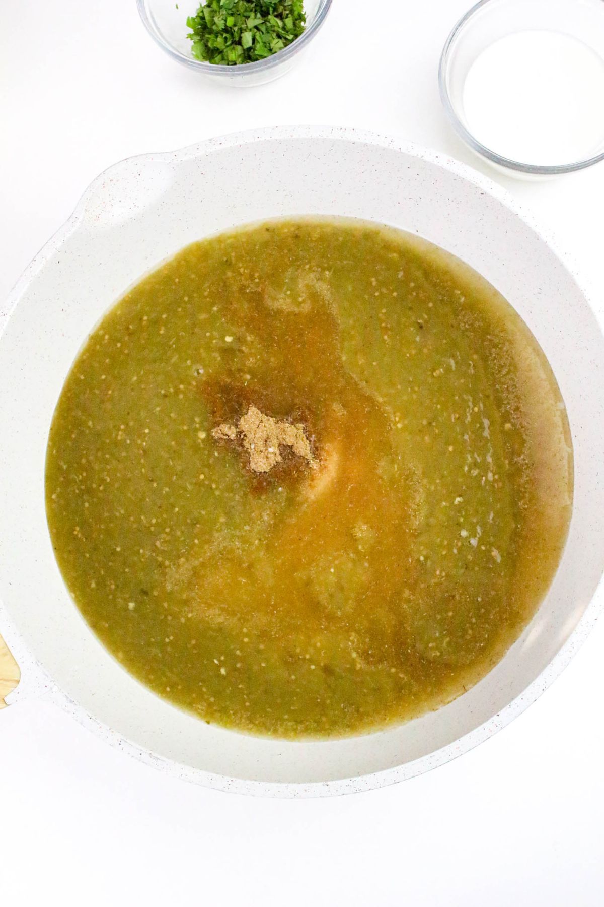 A white bowl filled with green sauce, likely salsa verde, with a pile of brown spices in the center. Small bowls with chopped herbs and a white ingredient are visible nearby on a white surface.
