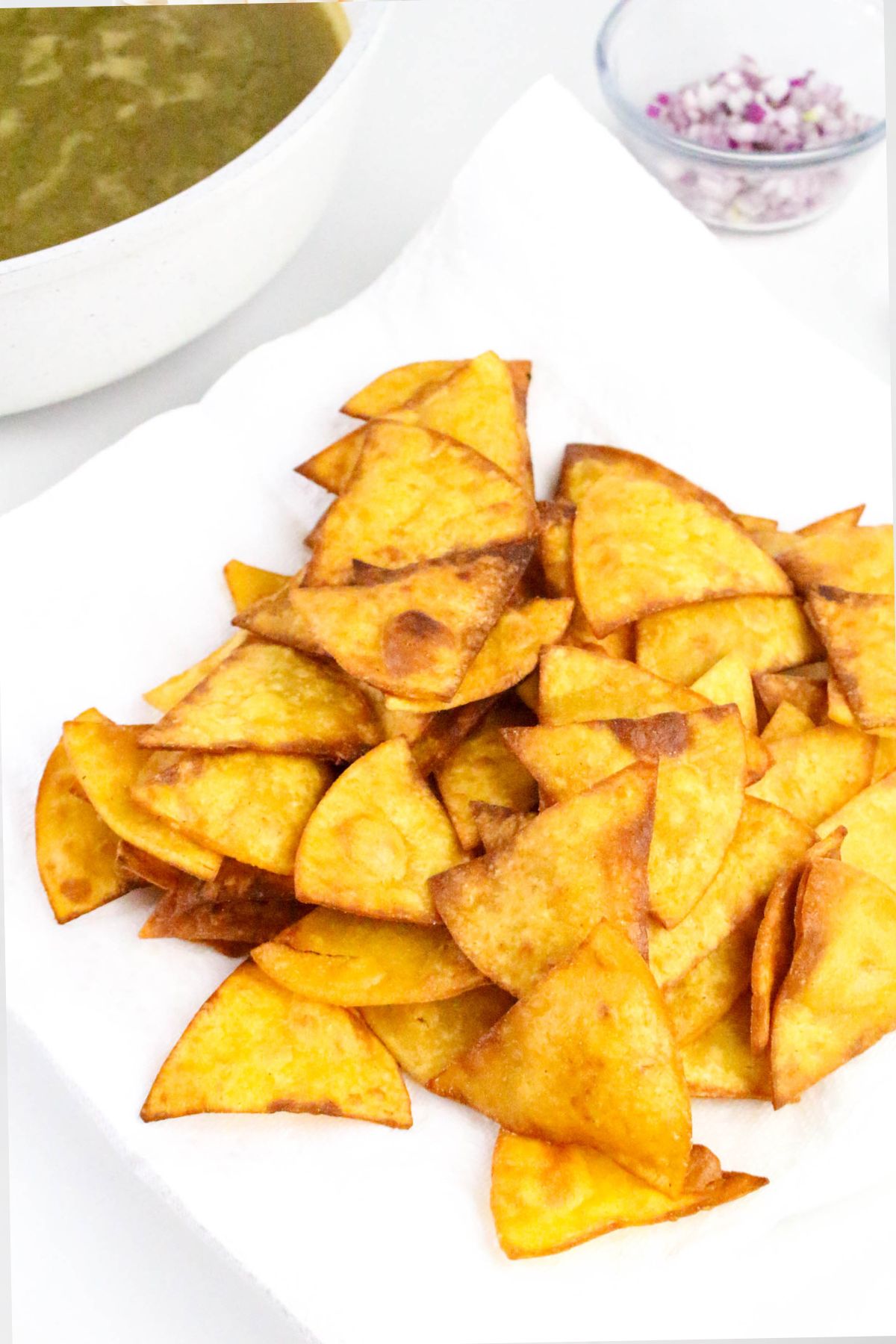 A pile of golden, triangle-shaped tortilla chips on a paper towel, with a bowl of green sauce and a small bowl of chopped red onions in the background.