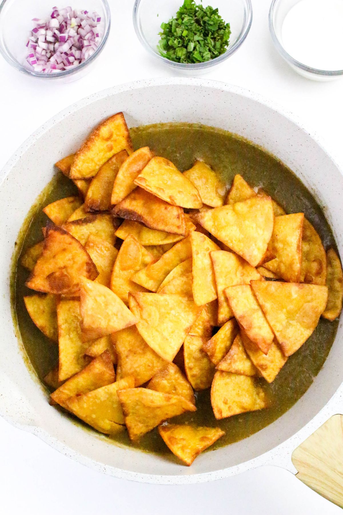 A white skillet contains crispy fried tortilla chips partially covered in green sauce. Nearby, small glass bowls hold chopped red onion, chopped cilantro, and a creamy white sauce.