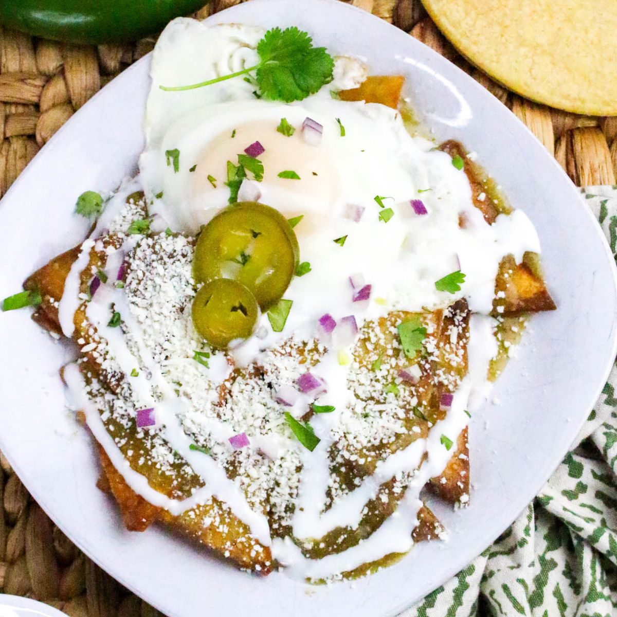 A plate of chilaquiles topped with a fried egg, jalapeño slices, crumbled cheese, diced red onion, cilantro, and a drizzle of crema, served on a white plate.