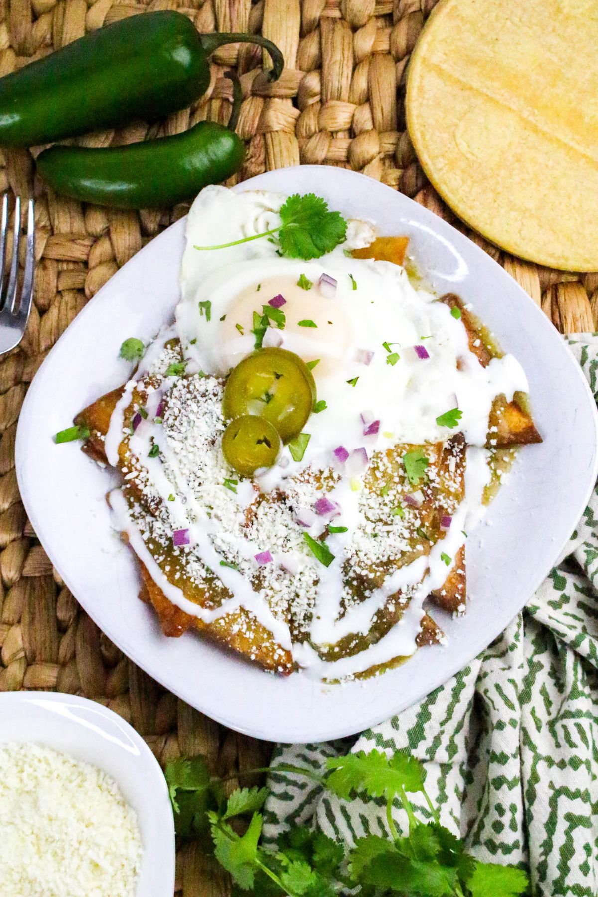 A white plate with chilaquiles topped with a fried egg, pickled jalapeños, sour cream, crumbled cheese, chopped red onion, and cilantro, set on a woven placemat next to corn tortillas and green peppers.