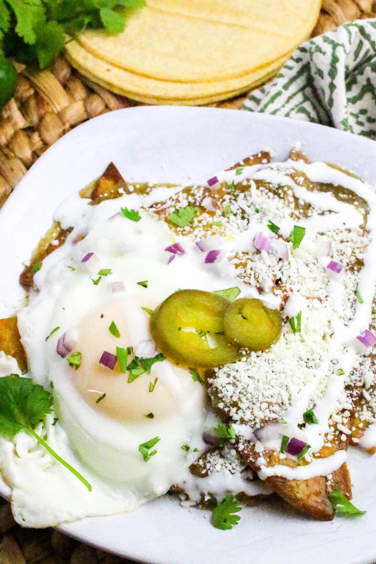 A plate of chilaquiles topped with a fried egg, sour cream, crumbled cheese, sliced jalapeños, chopped red onion, and cilantro. A stack of corn tortillas is visible in the background.
