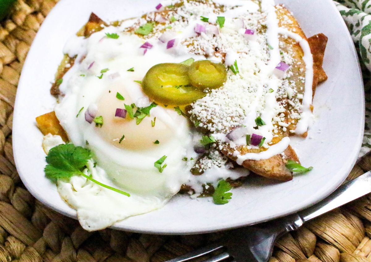 A plate of chilaquiles topped with a fried egg, sliced jalapeños, red onion, crumbled cheese, sour cream, and chopped cilantro. A fork and knife are placed next to the plate on a woven placemat.