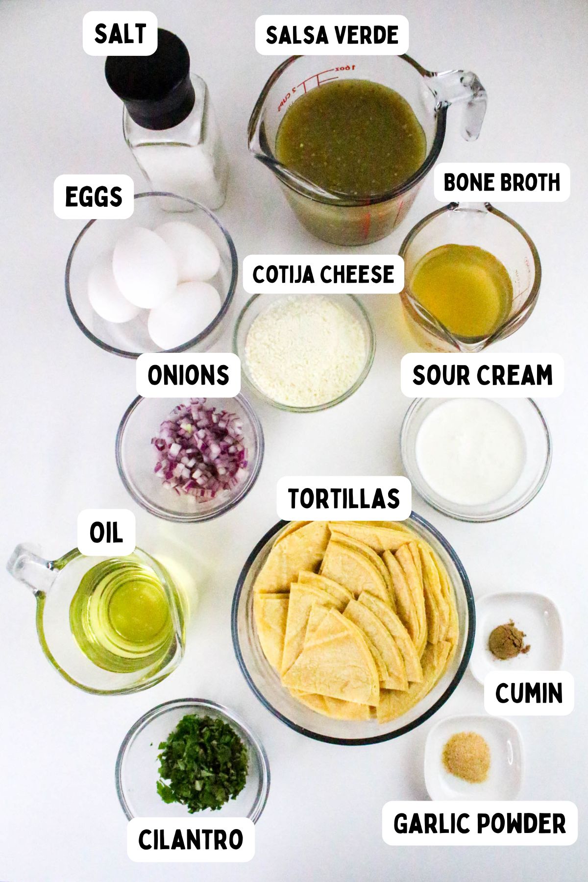 A set of labeled ingredients on a white surface, including salt, salsa verde, eggs, bone broth, cotija cheese, sour cream, chopped onions, oil, tortilla pieces, cumin, garlic powder, and chopped cilantro.