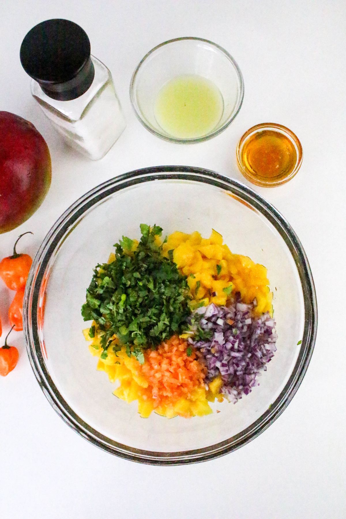 A glass bowl contains chopped cilantro, red onion, orange pepper, and mango. Surrounding the bowl are a mango, two small peppers, a bottle of salt, a small bowl of lime juice, and a small bowl of honey on a white surface.