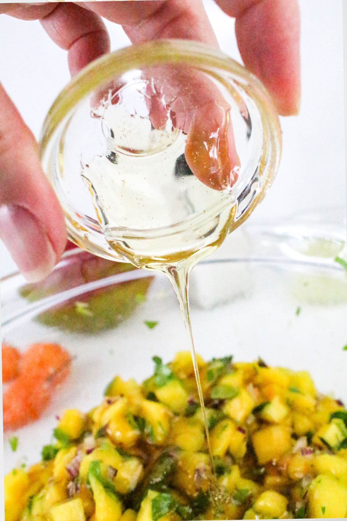 A close-up of a hand pouring honey from a small glass bowl onto a mixture of diced mango, red onion, and herbs in a larger glass bowl. Some other chopped ingredients are visible in the background.