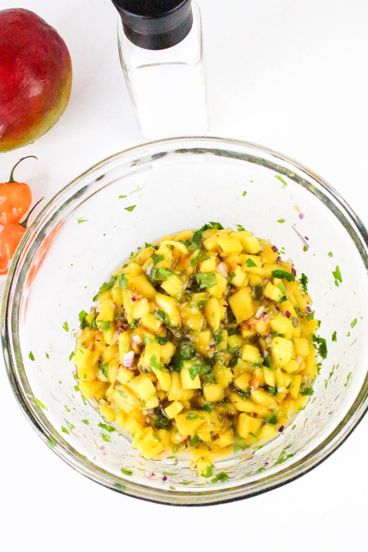 A glass bowl filled with a chopped mango salsa mixture sits on a white surface, surrounded by a whole mango, a habanero pepper, and a pepper grinder.