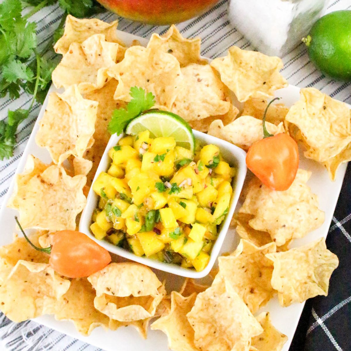 A square white plate with tortilla chips arranged around a bowl of mango salsa, garnished with a lime slice and cilantro. Two orange habanero peppers are placed on the plate beside the salsa.