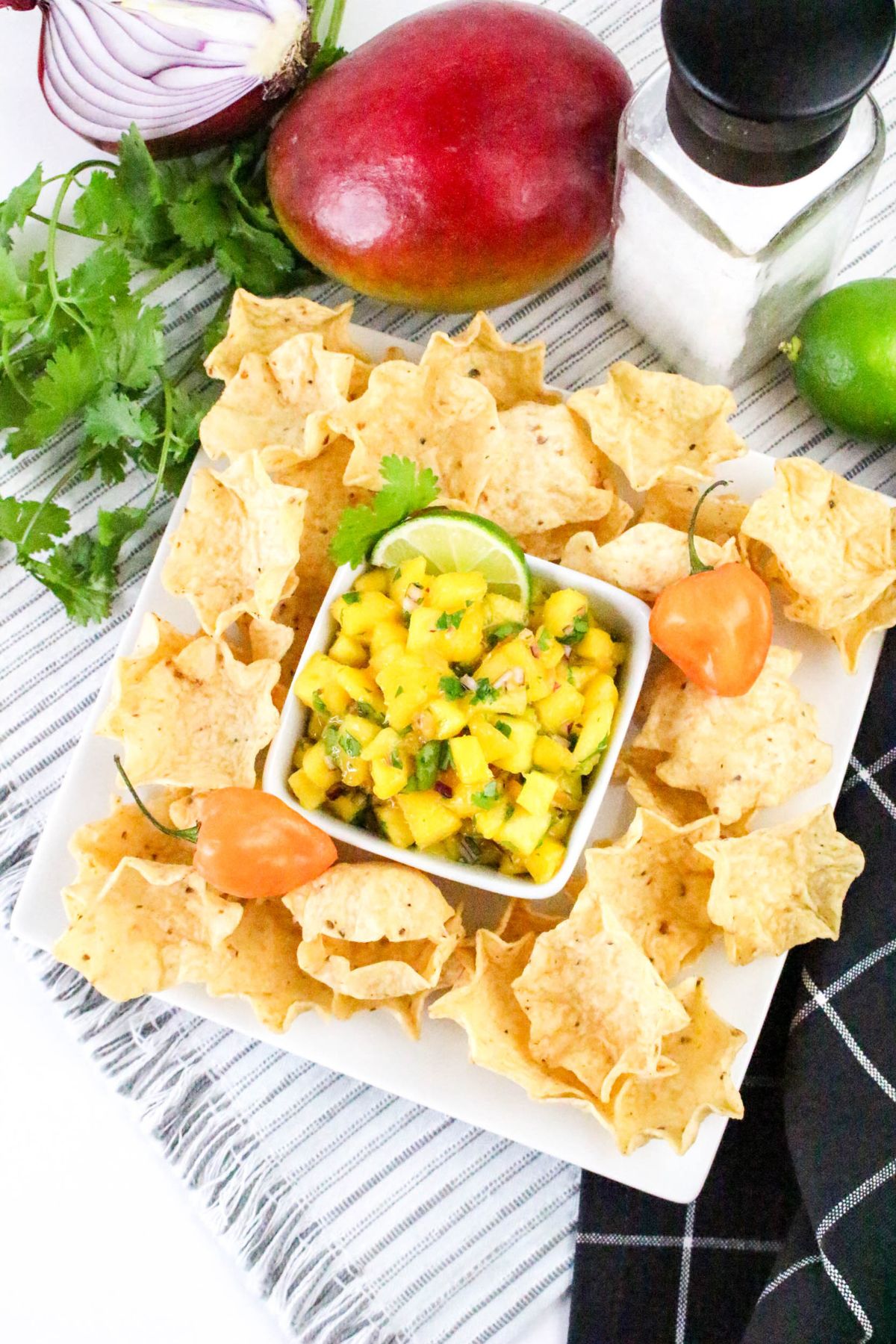 A square white plate holds tortilla chips arranged around a bowl of mango salsa, garnished with lime and cilantro. Nearby are a mango, habanero peppers, cilantro, a halved red onion, salt, and a lime on a striped cloth.