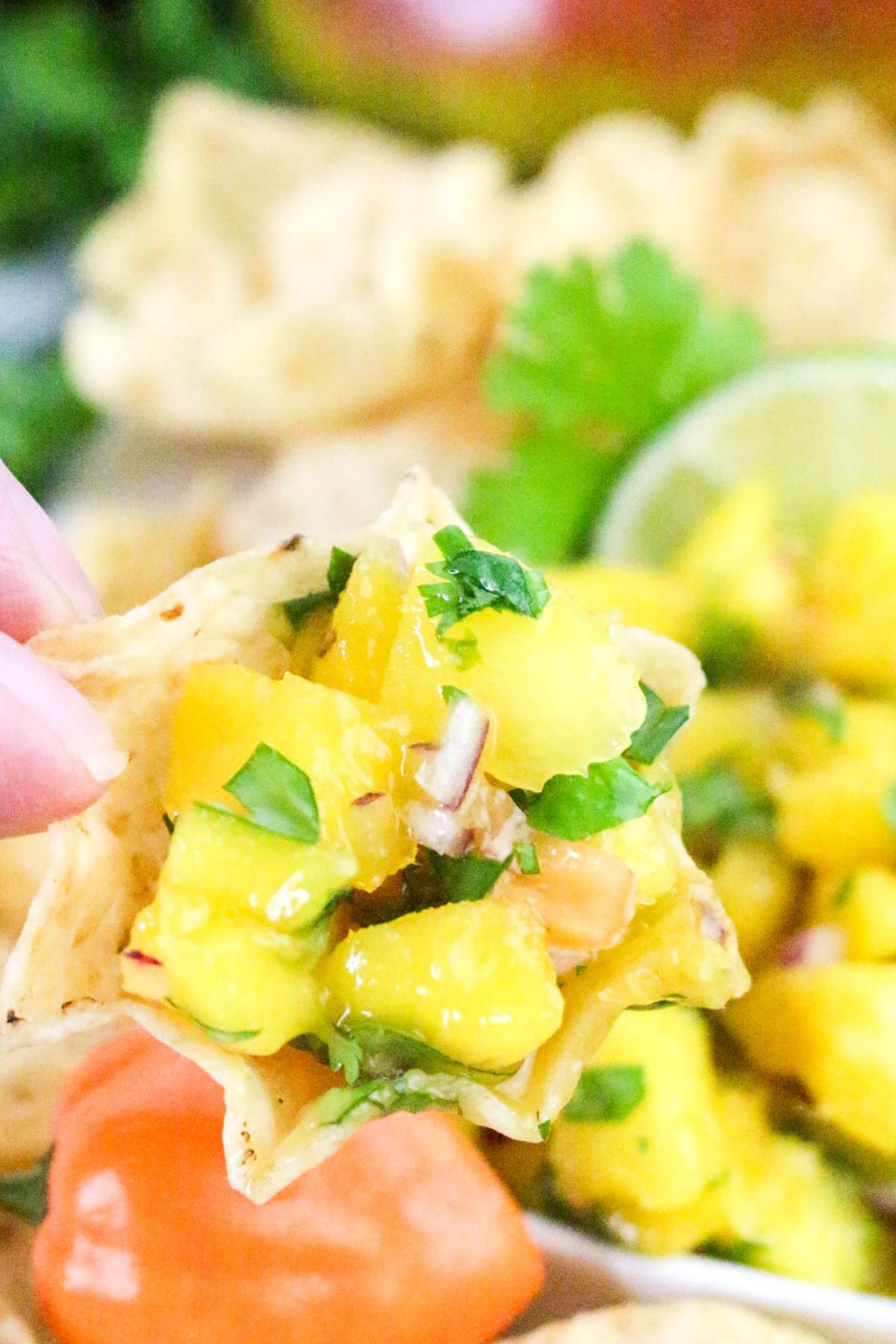 A close-up of a tortilla chip topped with mango salsa, showing diced mango, red onion, cilantro, and other ingredients. Other chips, salsa, and a lime are visible in the blurred background.