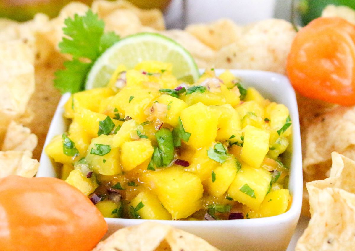 A square white bowl filled with mango salsa containing chopped mango, cilantro, and red onion, surrounded by tortilla chips, a lime slice, a sprig of cilantro, and orange peppers.