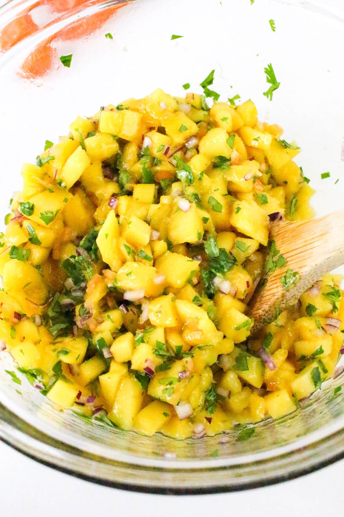 A glass bowl filled with mango salsa made of diced mango, chopped red onion, cilantro, and herbs, being mixed with a wooden spoon.