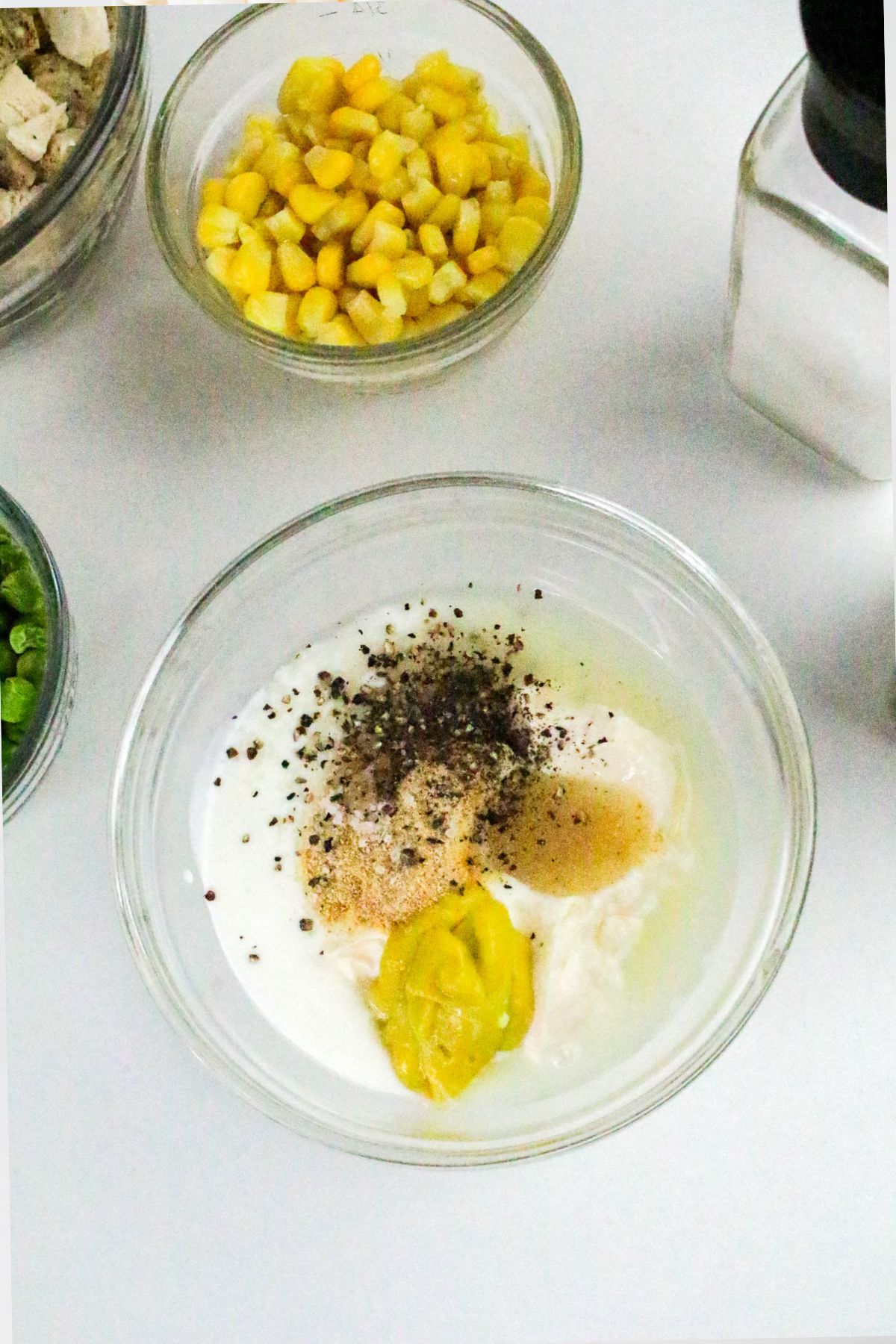 A glass bowl with mayonnaise, mustard, ground pepper, and seasoning next to a bowl of corn and another bowl with green beans on a white surface. A pepper grinder is partially visible in the top right.