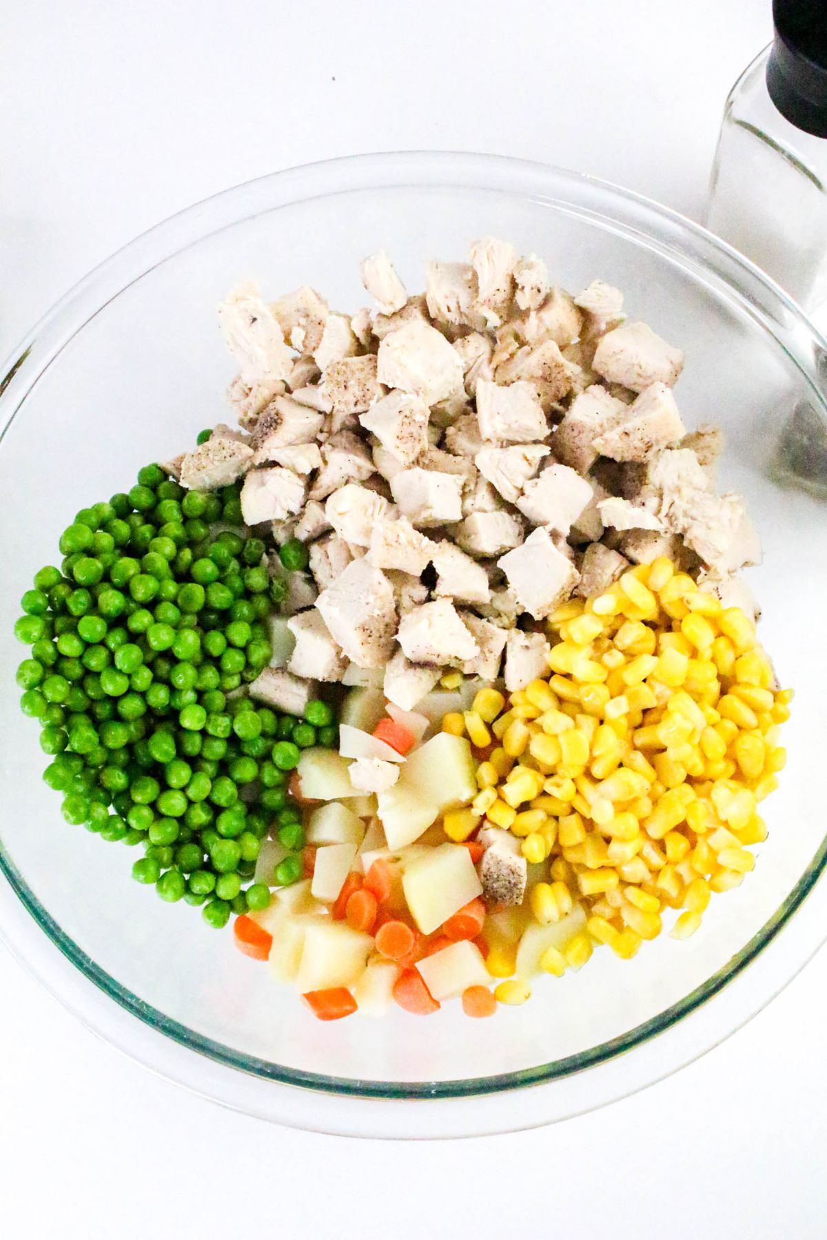 A glass bowl containing four separate sections of chopped cooked chicken, green peas, corn kernels, and a mix of diced carrots and potatoes, all on a white surface.