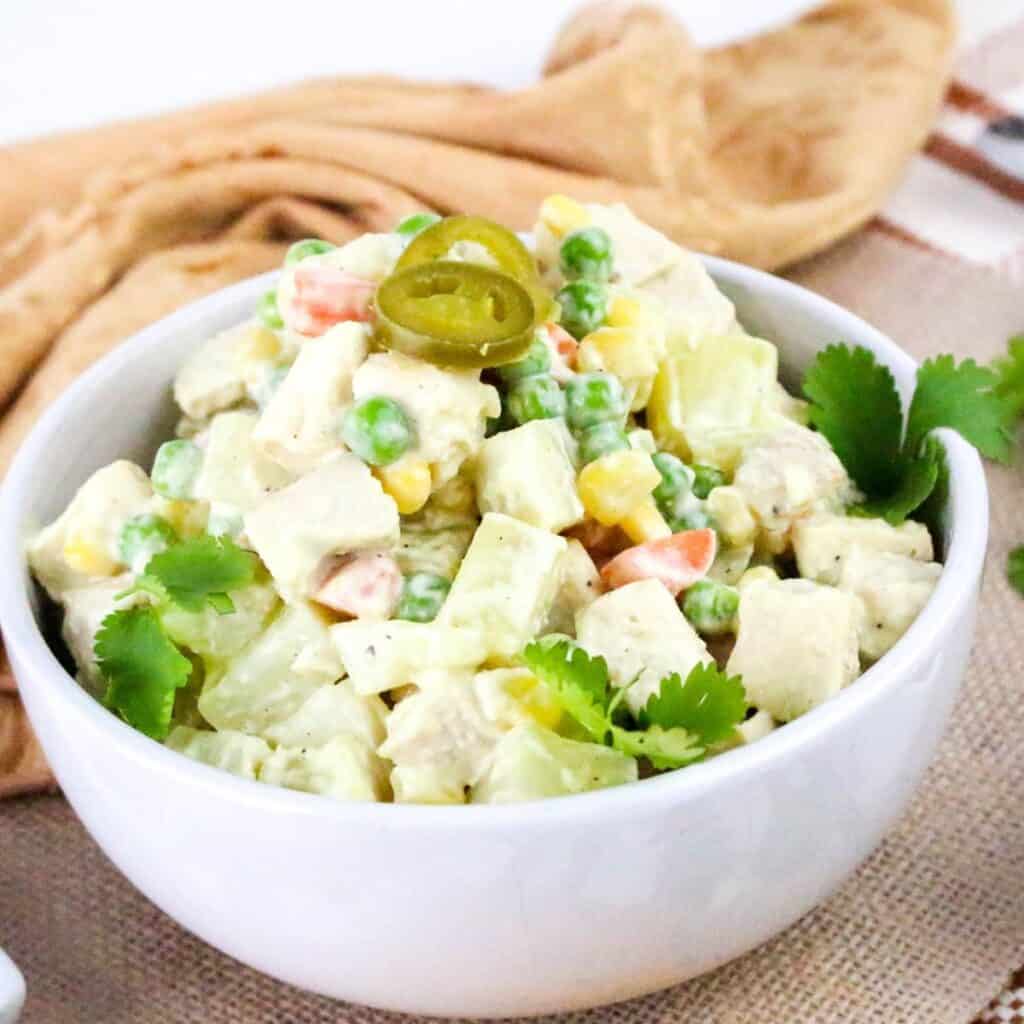 A white bowl filled with a mixed salad containing diced chicken, peas, corn, carrots, and potatoes in a creamy dressing, garnished with cilantro leaves and sliced jalapeño peppers. A tan cloth is in the background.