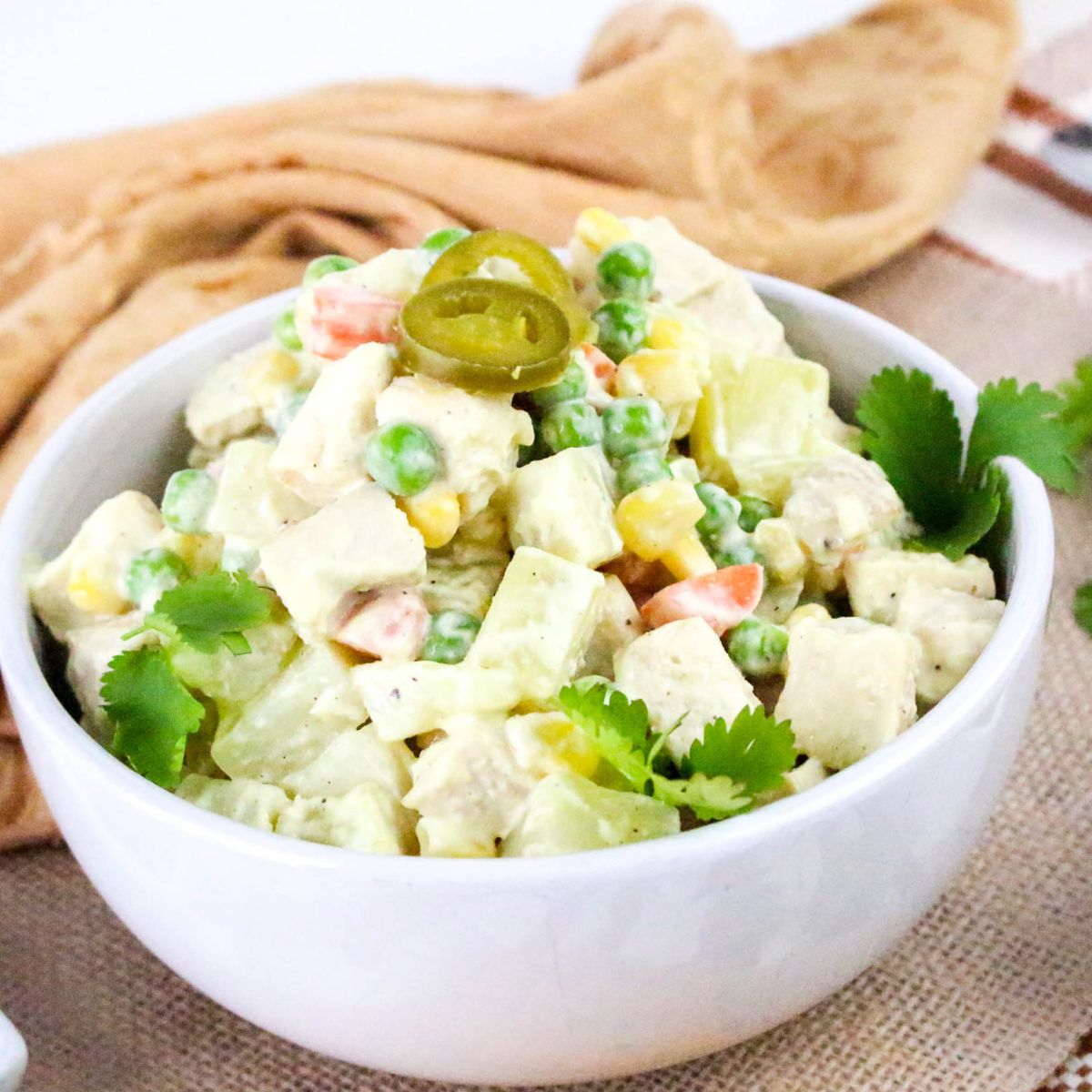 A white bowl filled with a mixed salad containing diced chicken, peas, corn, carrots, and potatoes in a creamy dressing, garnished with cilantro leaves and sliced jalapeño peppers. A tan cloth is in the background.