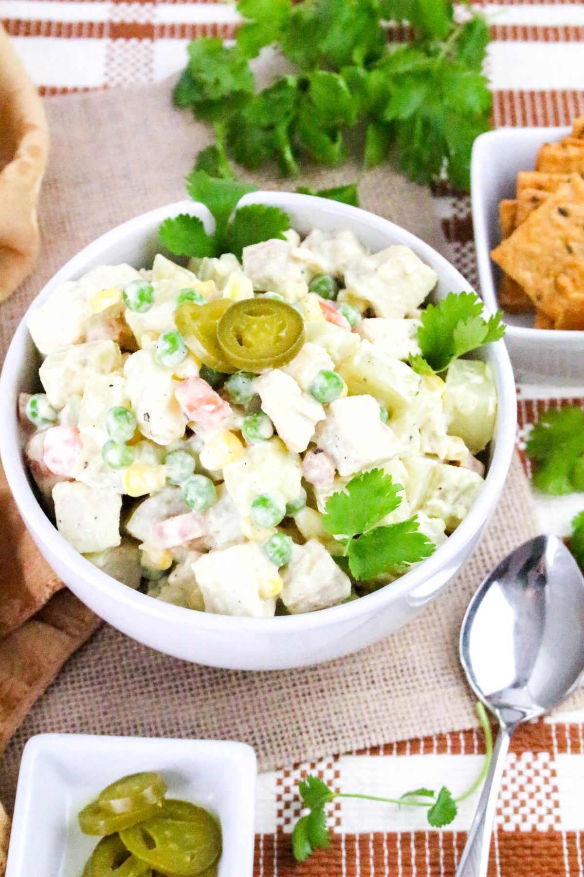 A white bowl filled with creamy potato salad with peas, carrots, and corn, garnished with cilantro and sliced jalapeños. The bowl is set on a cloth placemat near a spoon, square crackers, and extra jalapeño slices.