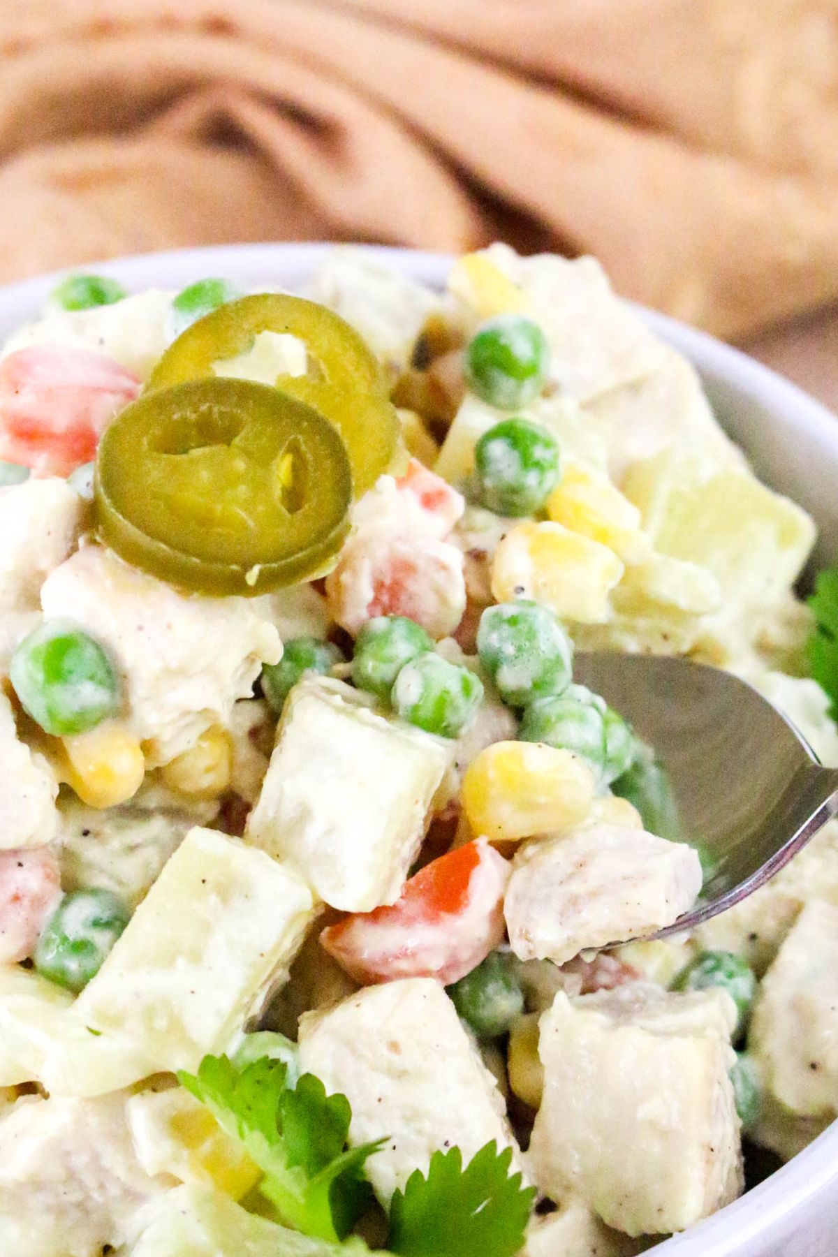 A close-up of a bowl of creamy chicken salad with diced chicken, peas, corn, carrots, and potato, garnished with sliced jalapeños and cilantro. A spoon is taking a portion from the bowl.