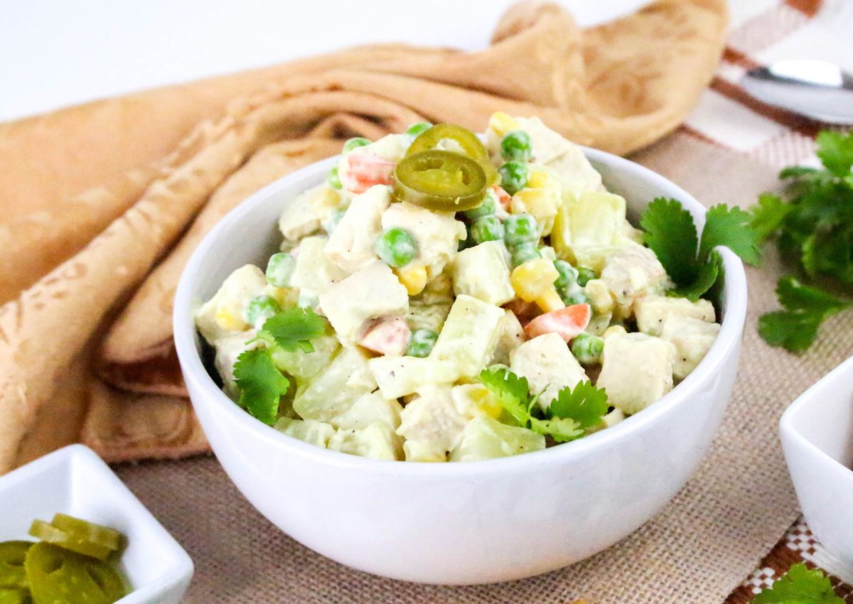 A white bowl filled with a mixed salad containing diced vegetables, possibly peas, carrots, corn, and cucumber, topped with sliced jalapeños and garnished with cilantro, placed on a table with a beige cloth.