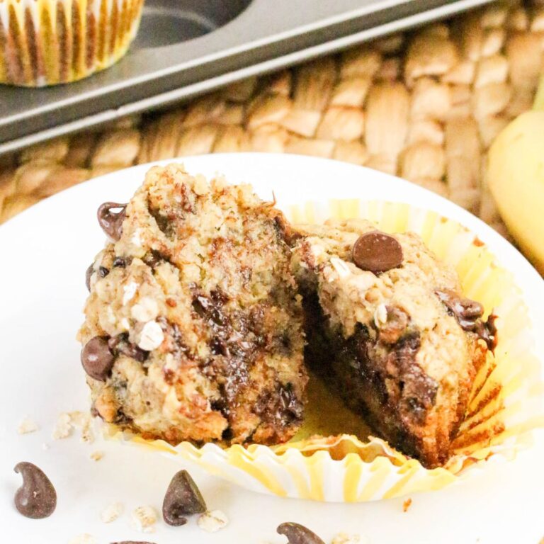 A banana chocolate chip muffin cut in half sits on a white plate, revealing a moist inside with melted chocolate chips. The muffin wrapper is partially peeled back, and a muffin tin is visible in the background.