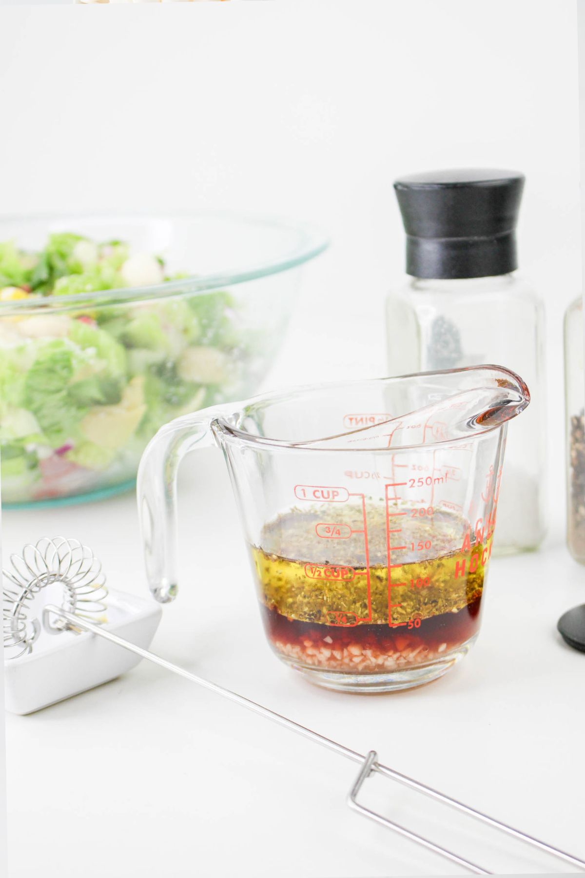 A glass measuring cup filled with salad dressing ingredients sits on a white surface. Nearby are a whisk, a bowl of salad, a salt shaker, and a pepper grinder.