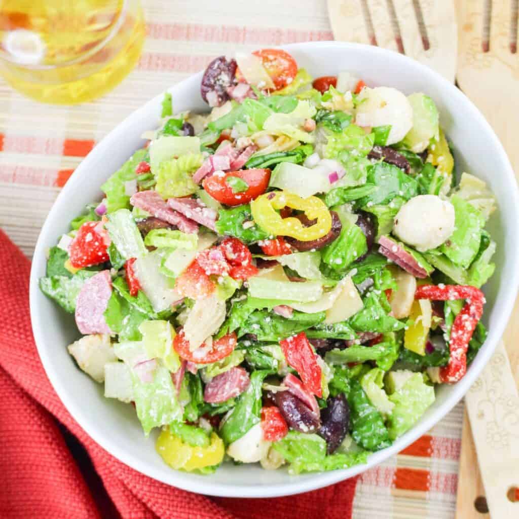 A white bowl filled with chopped salad including lettuce, olives, peppers, mozzarella balls, red onion, salami slices, and cherry tomatoes, placed on a red napkin with a drink and wooden utensils nearby.