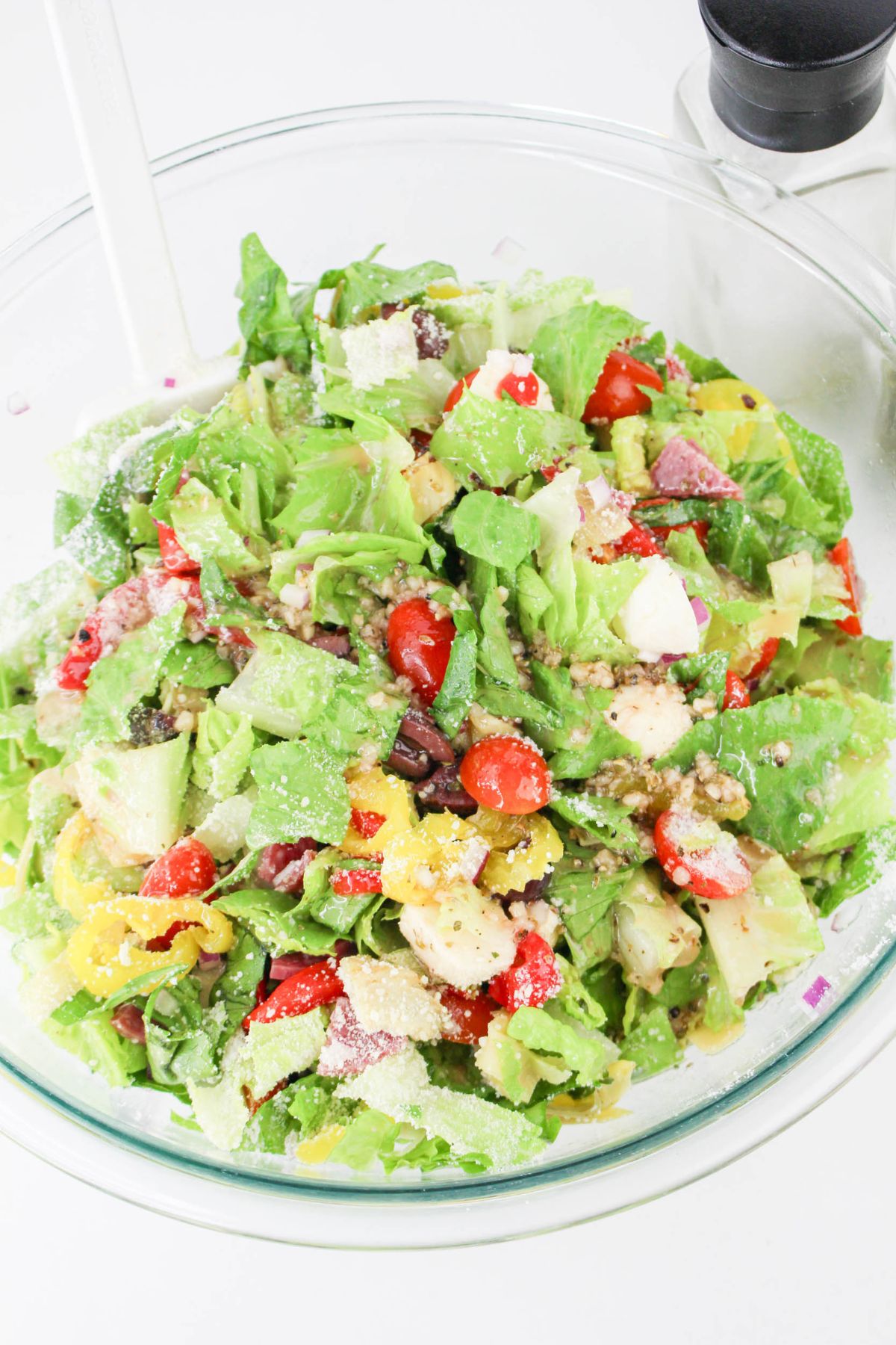 A clear glass bowl filled with chopped salad, including lettuce, cherry tomatoes, olives, pepperoncini, red onion, cheese, and topped with grated parmesan. A white serving utensil is in the bowl, and a pepper grinder is nearby.
