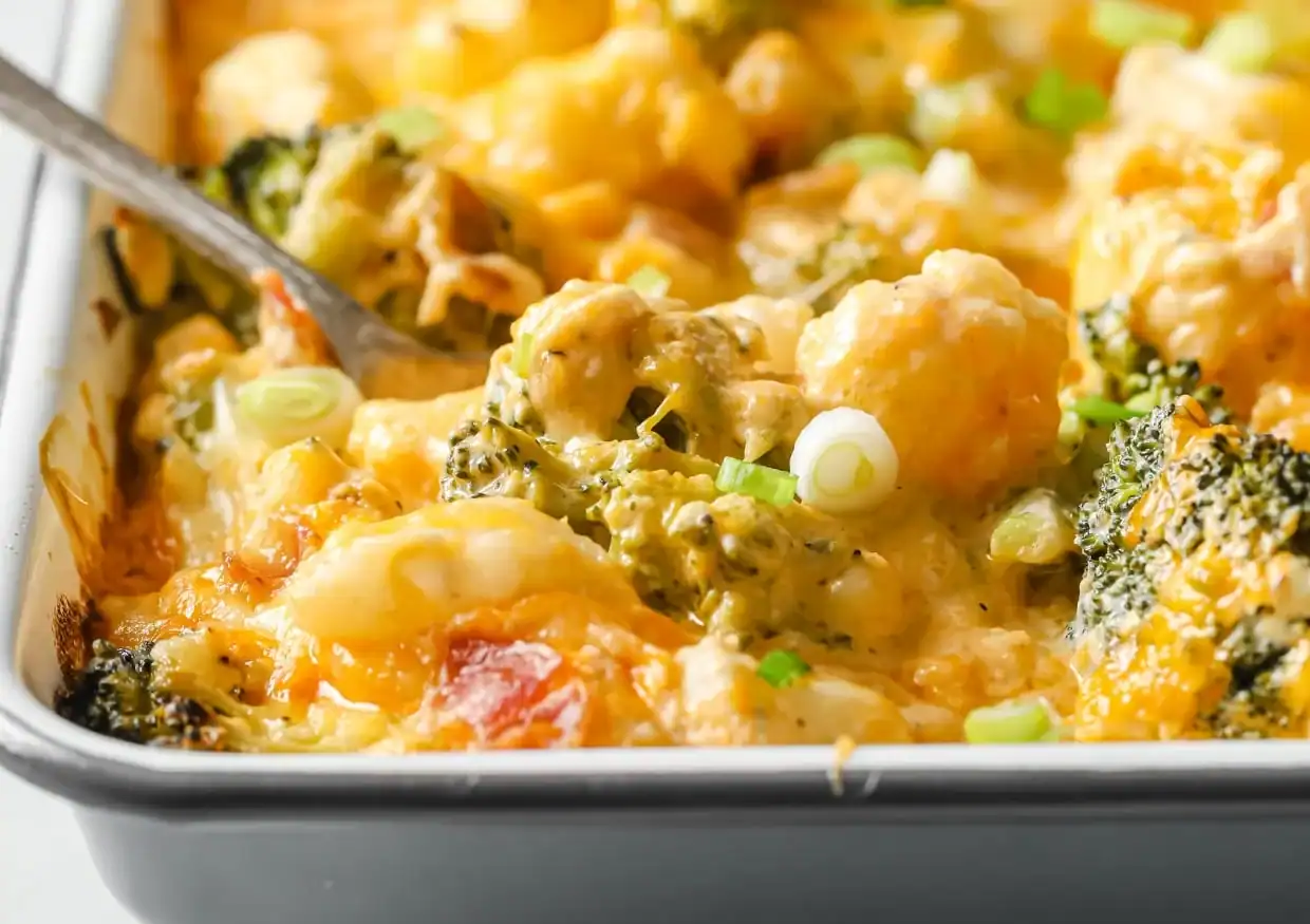 A close-up of a baked casserole with visible broccoli, cauliflower, melted cheese, and chopped green onions in a rectangular dish, with a serving spoon partially shown on the left.
