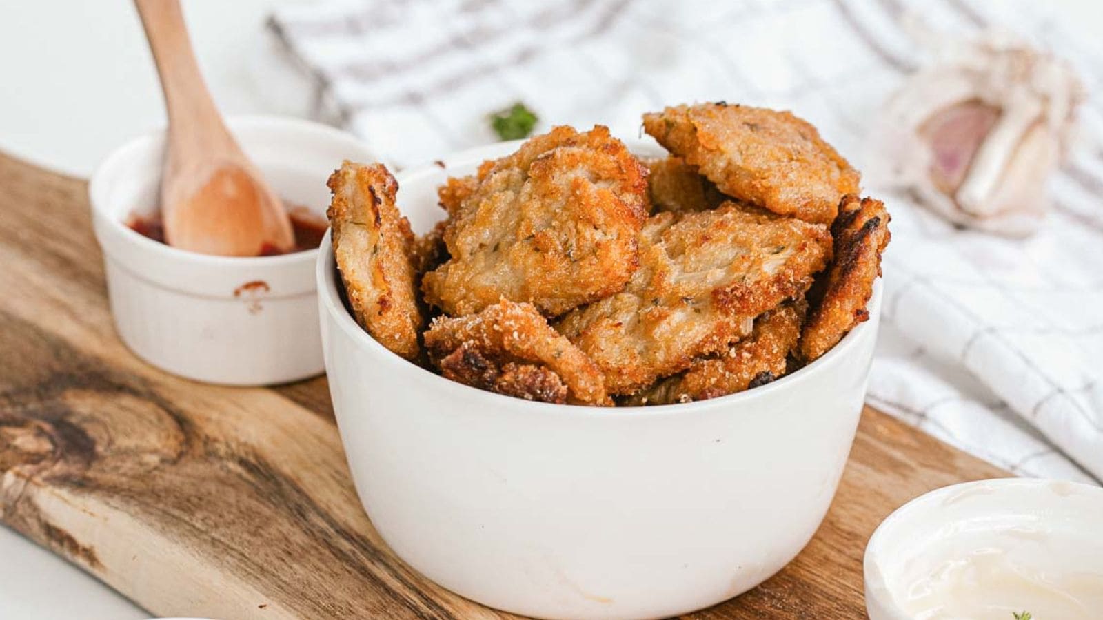 A white bowl filled with several pieces of crispy, golden-brown fried food sits on a wooden board next to a small dish of red sauce with a wooden spoon and a partial view of a white towel and garlic bulb in the background.