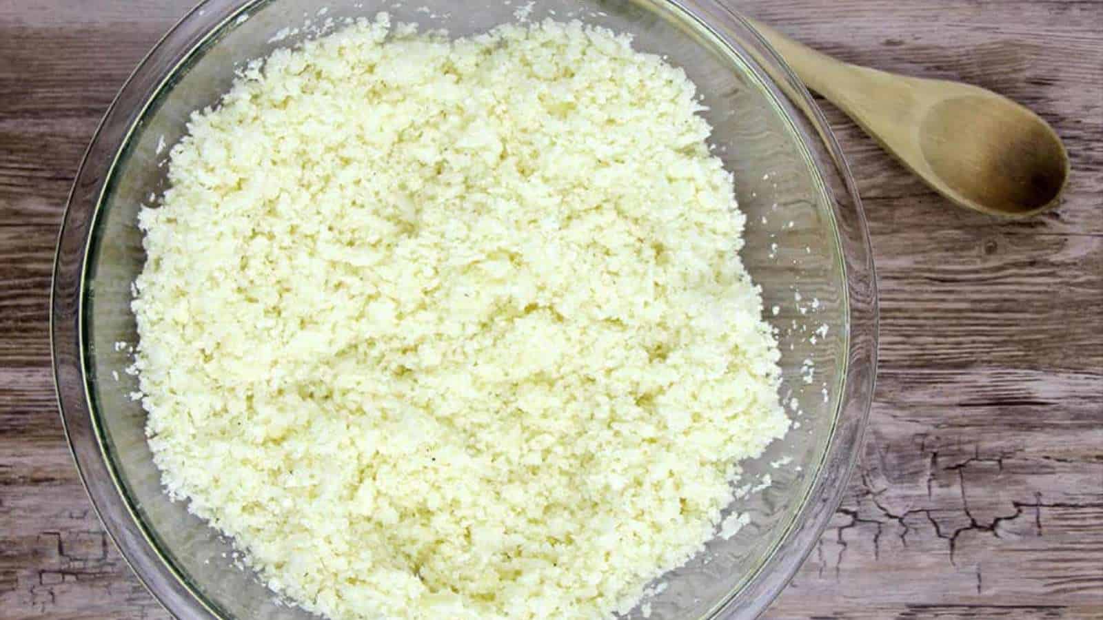 A clear glass bowl filled with riced cauliflower sits on a wooden surface next to a wooden spoon.