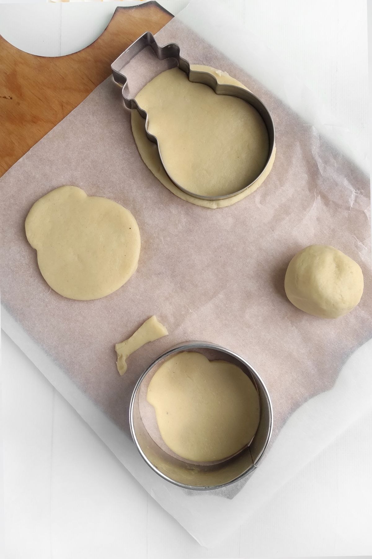 A baking sheet with parchment paper holds rolled Mrs Claus Cookies dough. Two metal cutters—one round and one snowman-shaped—are shown, along with cut dough shapes and a dough ball on a wooden surface.