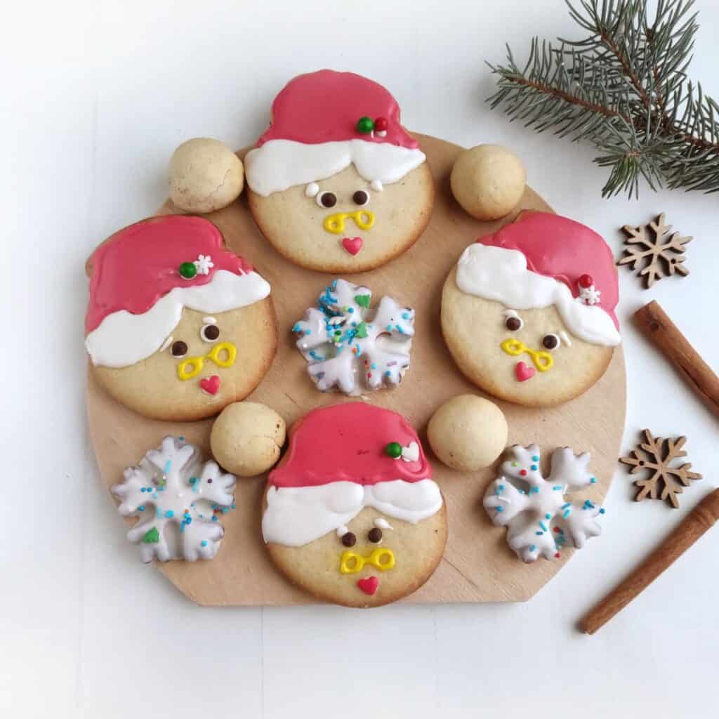 A round wooden board displays three Santa Claus cookies, two Mrs Claus Cookies, two snowflake cookies with blue sprinkles, three round cookies, cinnamon sticks, wooden snowflake ornaments, and a pine branch on a white surface.