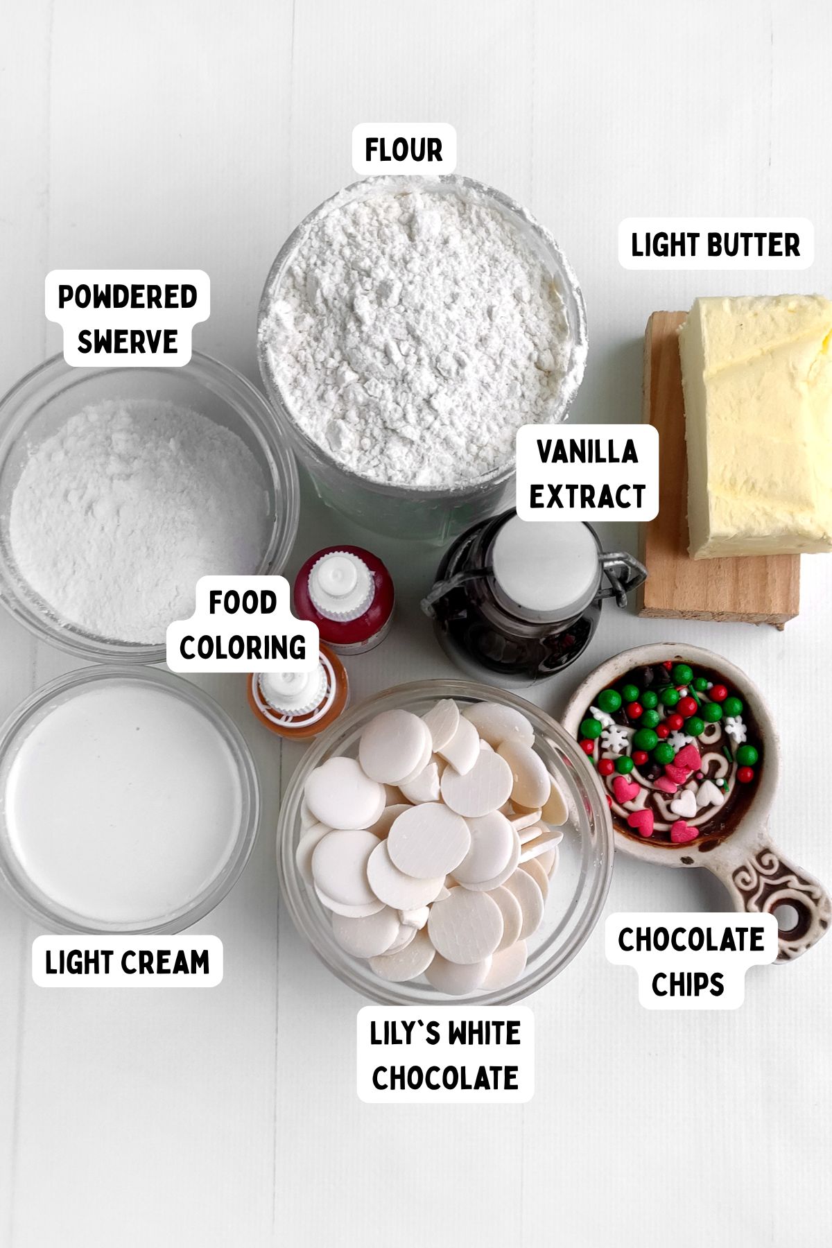 A top-down view of baking ingredients in bowls and jars, labeled as flour, light butter, vanilla extract, powdered Swerve, food coloring, light cream, Lily’s white chocolate, and chocolate chips for Mrs Claus Cookies.