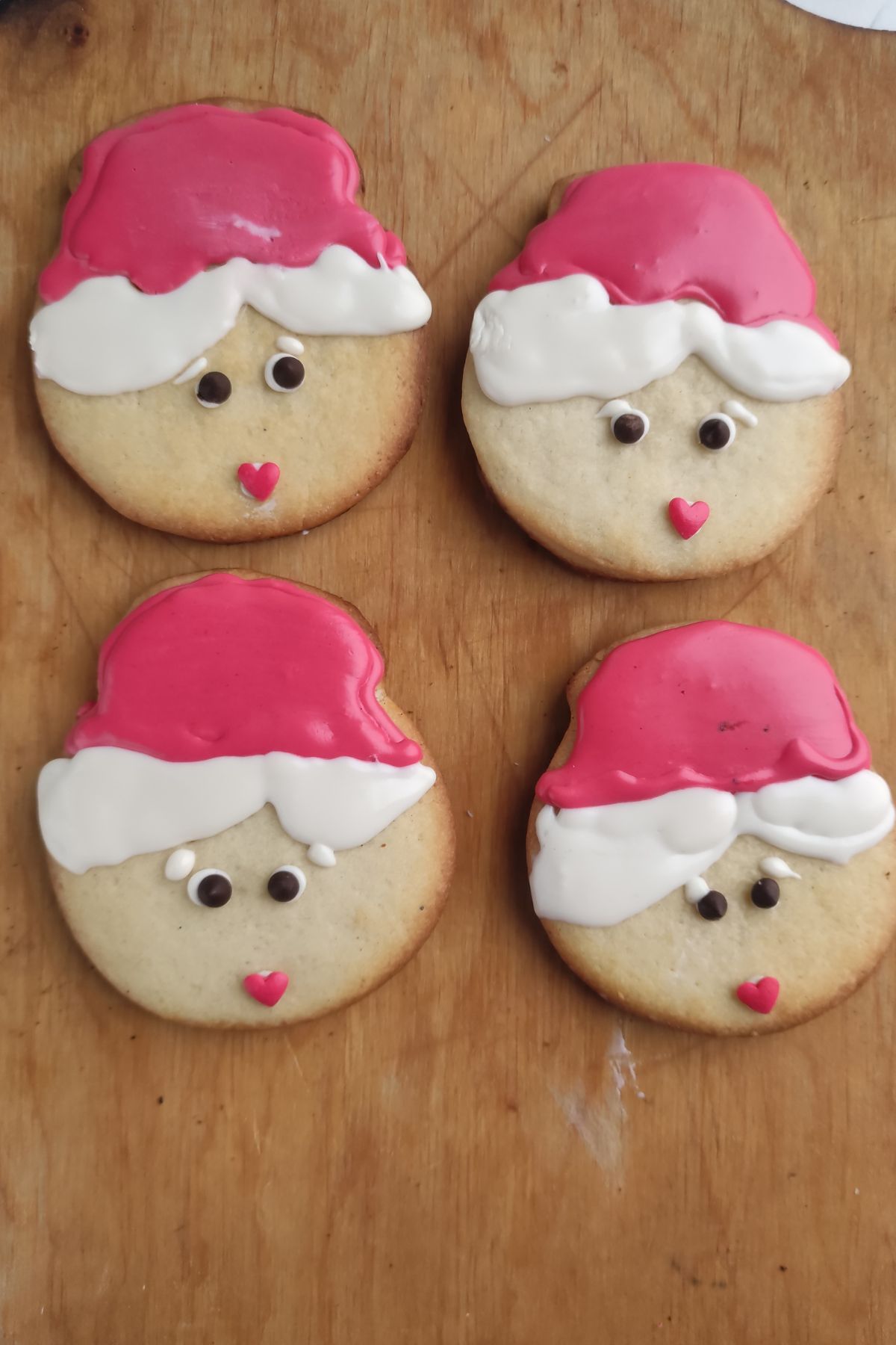 Four round Mrs Claus Cookies decorated with red and white icing hats, small candy eyes, and pink heart-shaped mouths, arranged on a wooden surface.