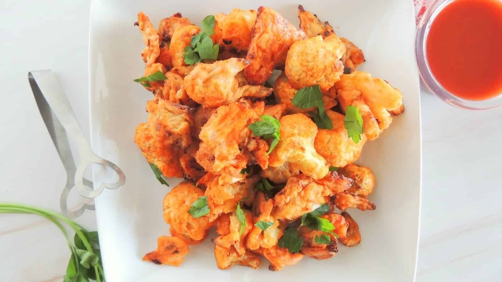 A white plate of baked buffalo cauliflower florets garnished with fresh parsley, next to a pair of metal tongs and a glass of red sauce on a light surface.