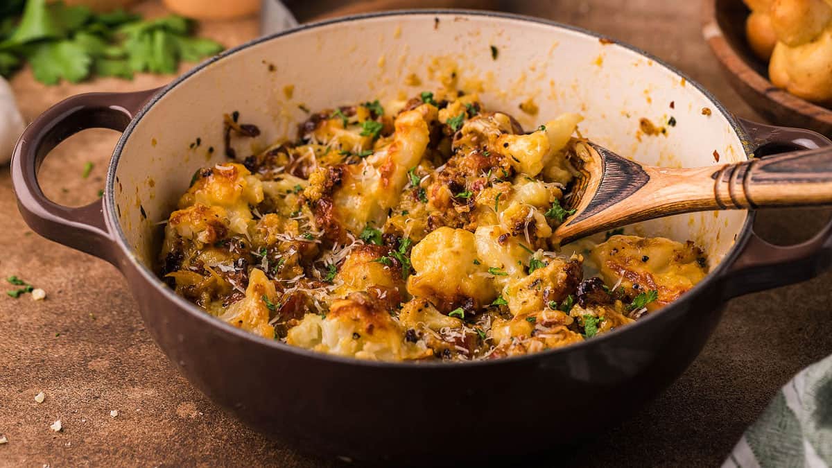 A brown ceramic pot filled with baked cauliflower, topped with melted cheese and herbs, sits on a table. A wooden spoon rests inside the pot, and there are scattered herbs around.
