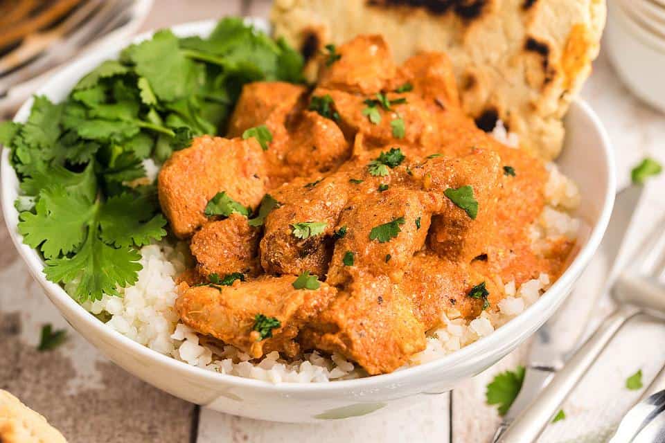 A white bowl filled with rice topped with pieces of chicken in orange sauce, garnished with chopped cilantro, and served with fresh cilantro leaves and a piece of flatbread.