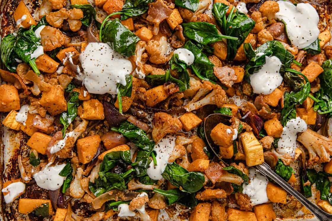 A sheet pan filled with roasted vegetables including sweet potatoes, cauliflower, spinach, and red onion, topped with dollops of white sauce and freshly ground black pepper. A serving spoon rests on the pan.