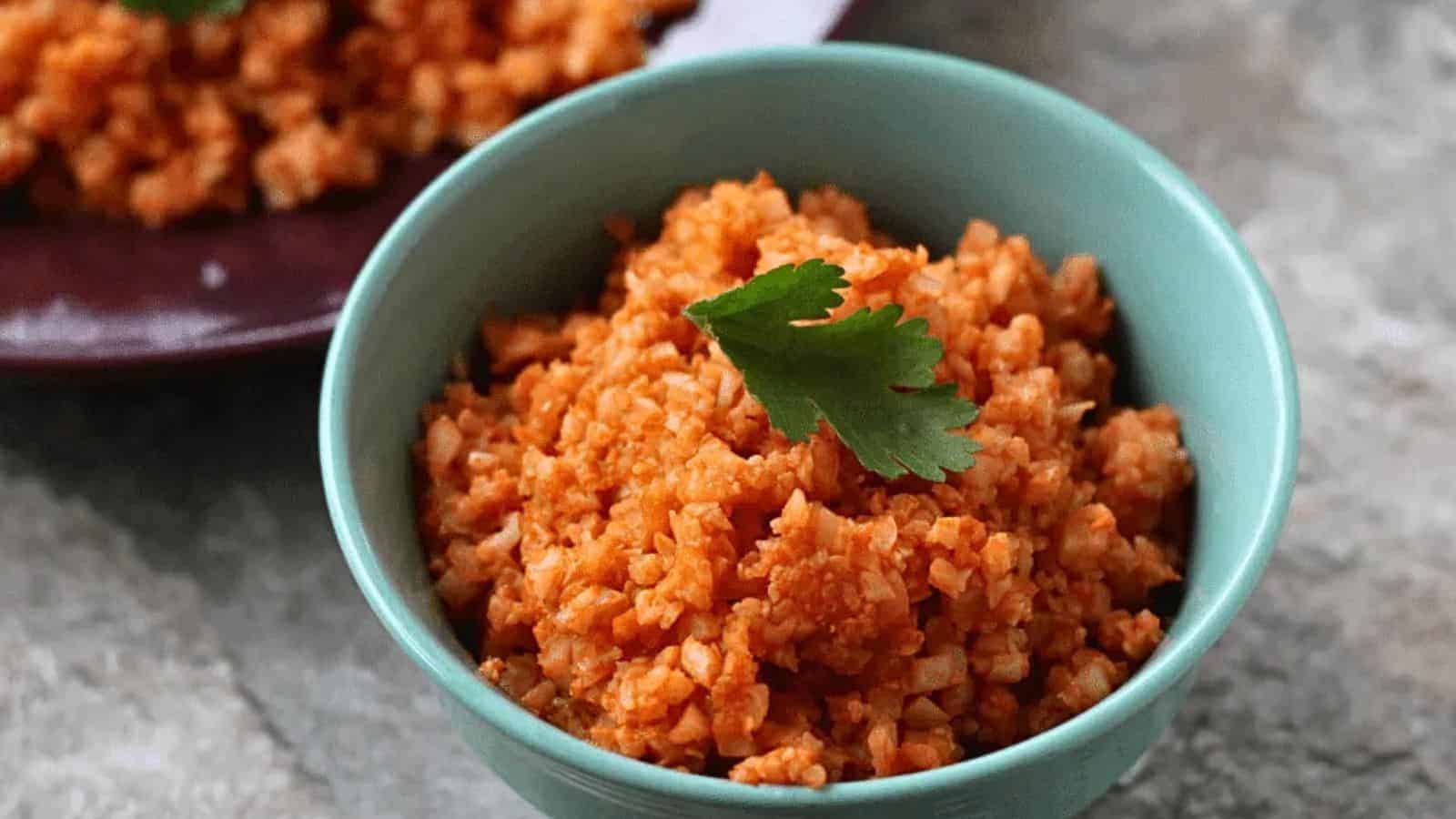 A light blue bowl filled with orange-colored rice, garnished with a sprig of cilantro. A plate with a similar rice dish is partially visible in the background. The surface underneath is gray and textured.