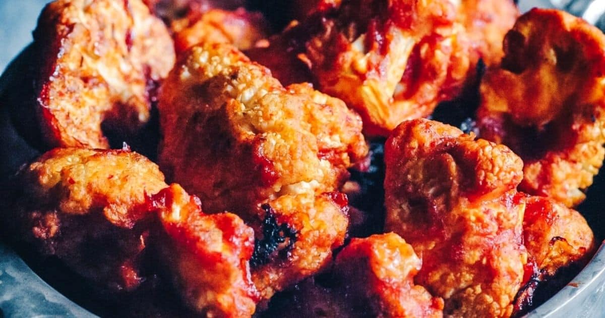 A close-up view of several pieces of buffalo cauliflower, coated in a red-orange sauce and slightly charred, arranged together on a plate.