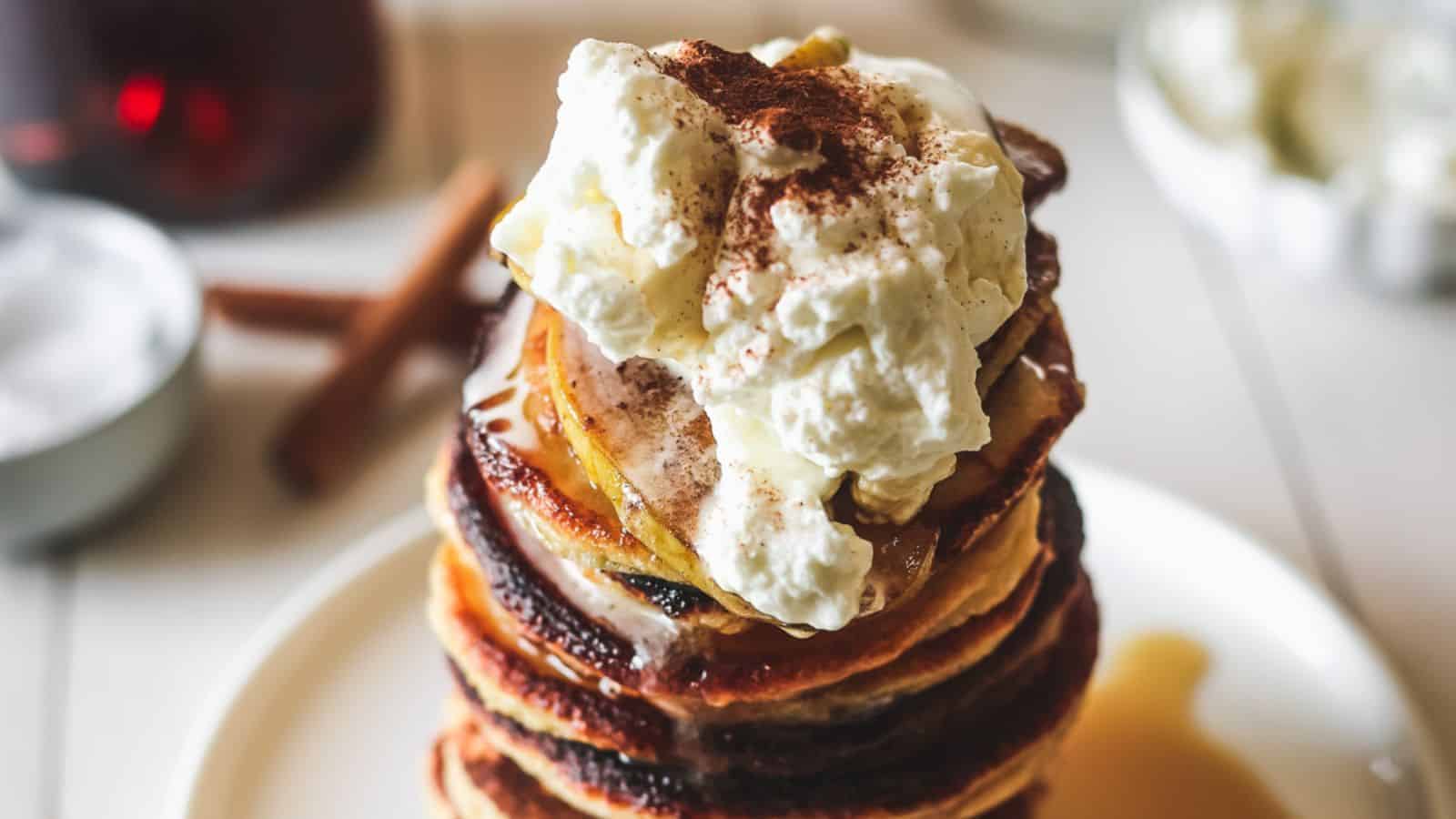 A stack of pancakes topped with a large dollop of whipped cream and a sprinkle of cinnamon, with syrup drizzling down the sides, on a white plate. Cinnamon sticks and a small bowl are in the background.