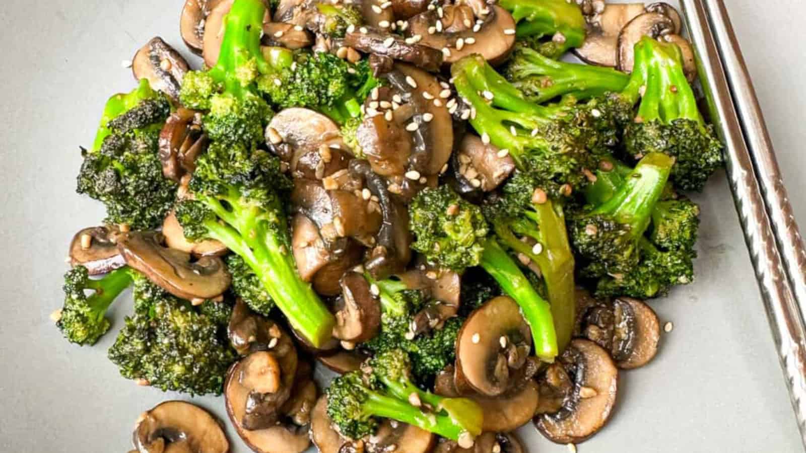 A plate of sautéed broccoli and sliced mushrooms garnished with sesame seeds, served with a pair of metal chopsticks on the side.