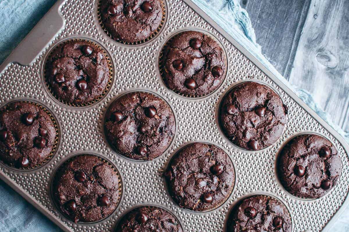 A muffin tin filled with twelve baked chocolate muffins, each topped with chocolate chips, sits on a textured surface with a blue cloth partially visible to the side.